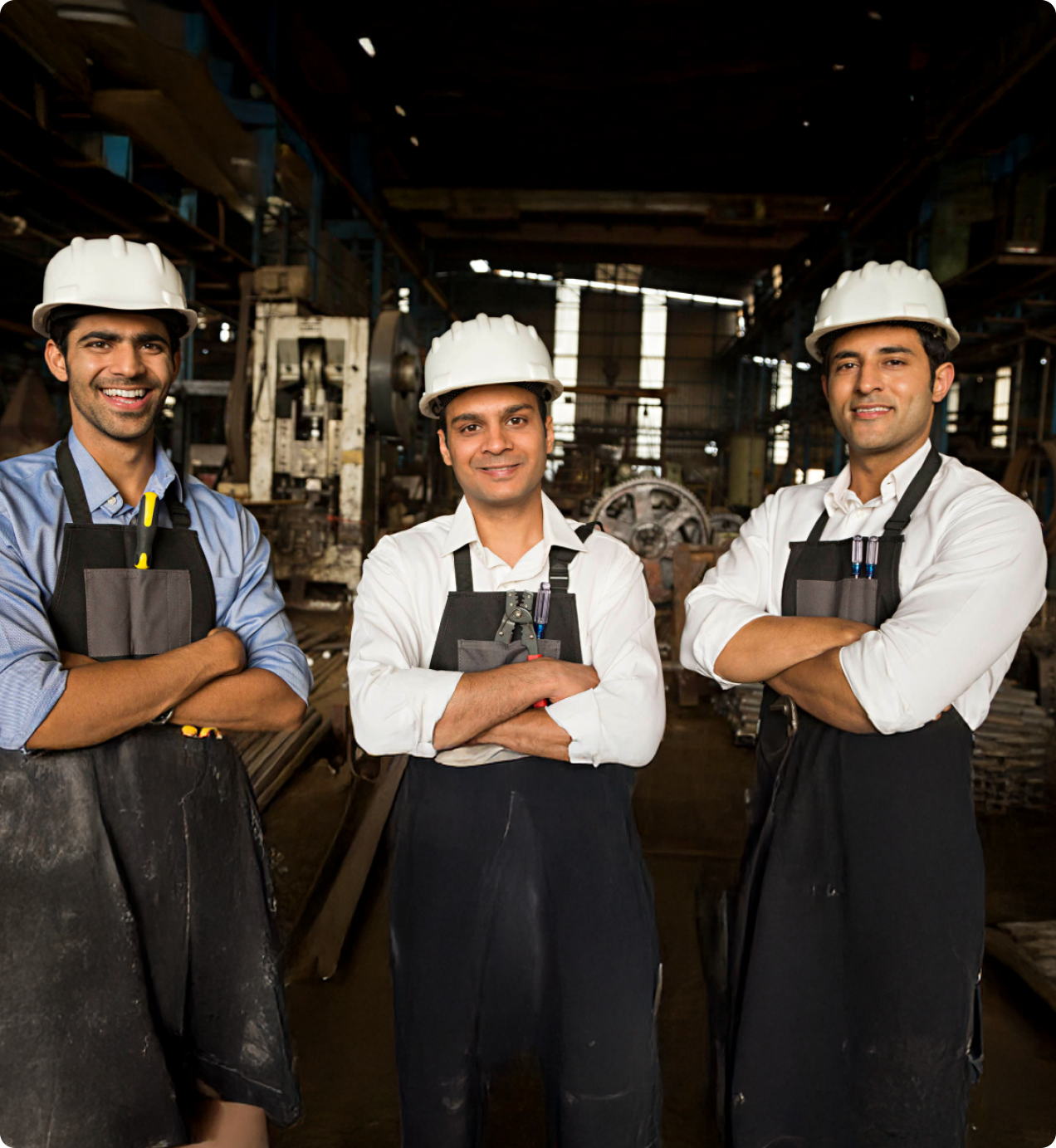 Three male factory workers wearing white safety helmets and black aprons standing with arms crossed inside an industrial workshop.
