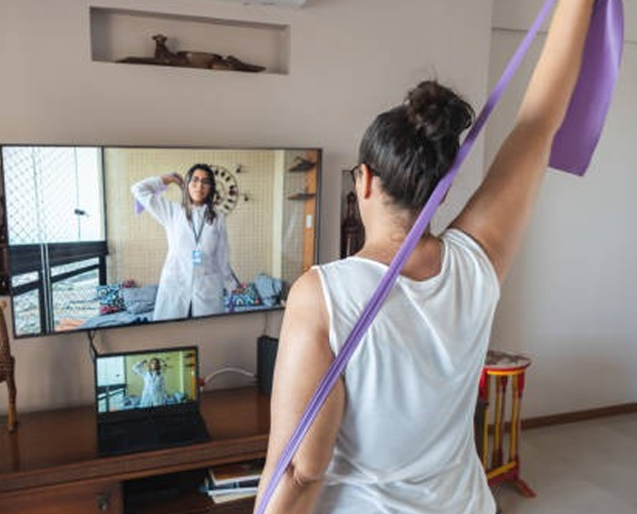 Person with hair in a bun doing resistance band exercises following a virtual trainer on a TV and laptop screen.